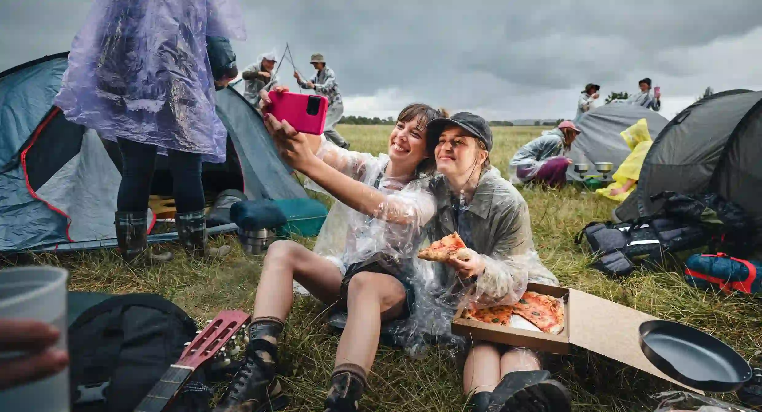Dvě kamarádky v pláštěnkách si na zablácené kempu dělají selfie s krabicí pizzy
